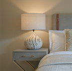Bedside table with a lamp and decorative items next to a bed in a bedroom setting.